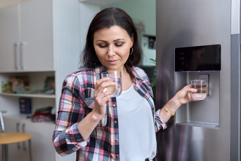 Water dispenser, woman taking cold water into glass from home refrigerator.